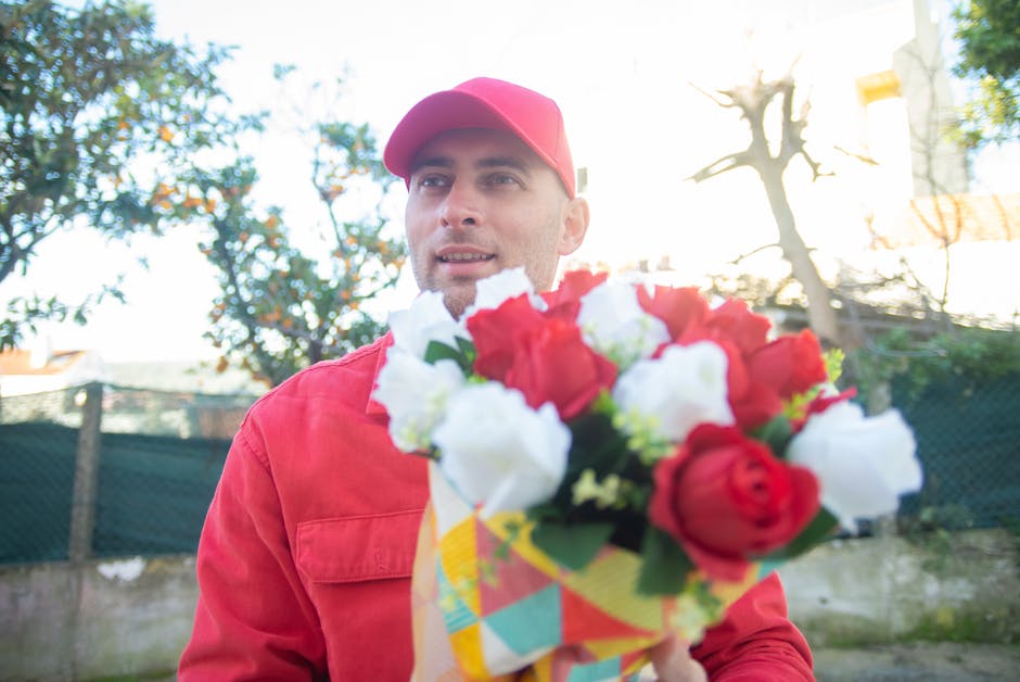 A delivery person wearing a red cap and matching red jacket holds a fresh floral bouquet in front of them during daylight. The bouquet features a mix of vibrant red and white roses with green foliage,